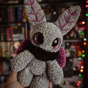 Puede incluir: Un peluche hecho a mano con forma de polilla, en tonos grises con detalles morados. La criatura tiene ojos grandes y brillantes y detalles en forma de hoja en las orejas. Cuenta con una bufanda marr&oacute;n oscuro y alas moradas.
