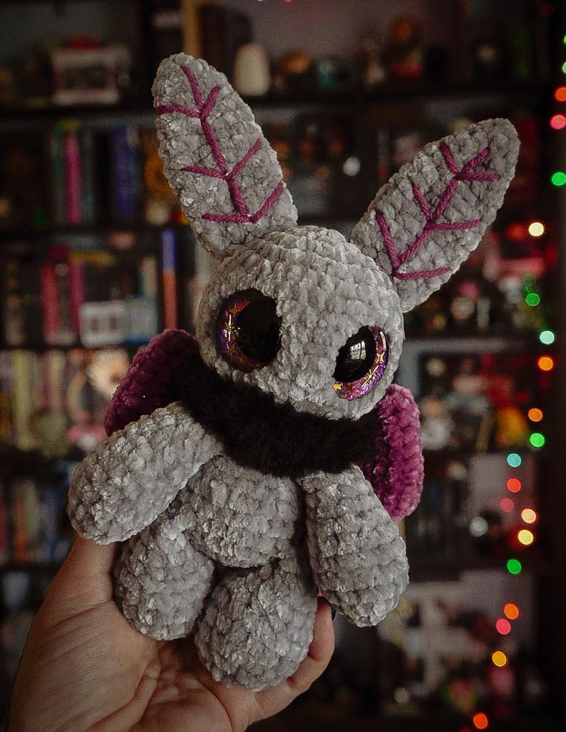 Puede incluir: Un peluche hecho a mano con forma de polilla, en tonos grises con detalles morados. La criatura tiene ojos grandes y brillantes y detalles en forma de hoja en las orejas. Cuenta con una bufanda marr&oacute;n oscuro y alas moradas.