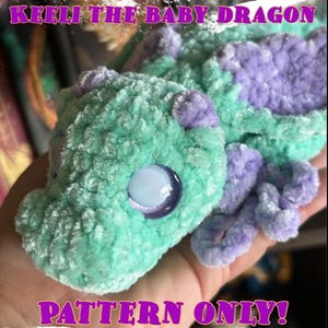 以下が含まれることがあります： 赤ちゃんドラゴンアミグルミの編み物パターン。ドラゴンは緑と紫で、大きくキラキラした目があります。パターンは販売されており、完成したおもちゃではありません。画像には「Keeli the Baby Dragon」と「Pattern Only！」のテキストが表示されています。