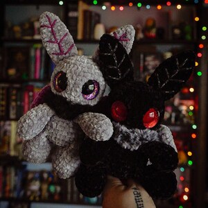 Puede incluir: Dos juguetes de peluche con forma de polilla. Uno es gris con detalles morados y ojos iridiscentes, el otro es negro con ojos rojos. Ambos tienen grandes orejas en forma de hoja. Se sostienen frente a un fondo de estanter&iacute;as y luces de colores.