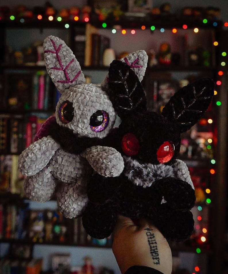 Puede incluir: Dos juguetes de peluche con forma de polilla. Uno es gris con detalles morados y ojos iridiscentes, el otro es negro con ojos rojos. Ambos tienen grandes orejas en forma de hoja. Se sostienen frente a un fondo de estanter&iacute;as y luces de colores.