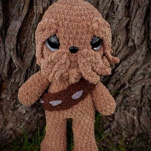 May include: A handmade brown crochet doll with large, expressive eyes and a detailed face. The doll has a brown belt with gray accents and is standing on green grass. The background features a textured tree trunk.