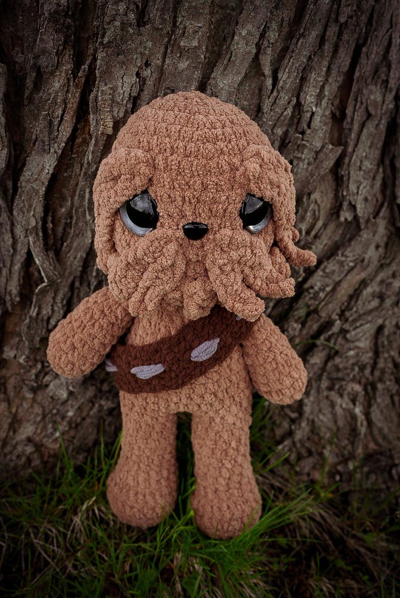 May include: A handmade brown crochet doll with large, expressive eyes and a detailed face. The doll has a brown belt with gray accents and is standing on green grass. The background features a textured tree trunk.