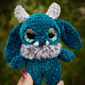May include: A teal plush monster with large, glossy black eyes and teal accents. It has grey horns and a fluffy grey collar. The handmade creature is soft and cuddly, perfect for a child's toy or a unique decorative item.