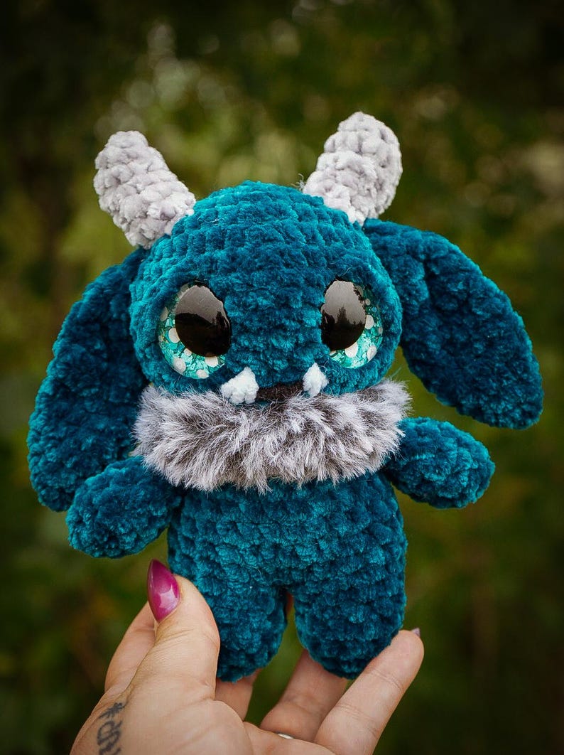May include: A teal plush monster with large, glossy black eyes and teal accents. It has grey horns and a fluffy grey collar. The handmade creature is soft and cuddly, perfect for a child's toy or a unique decorative item.