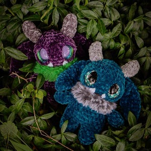 May include: Two plush monster toys nestled amongst green leaves. One is purple with green and grey accents, the other is teal with grey accents. Both have large, glittery eyes and faux fur details.