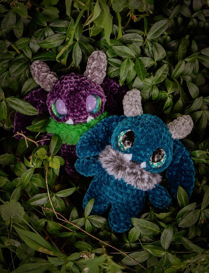 May include: Two plush monster toys nestled amongst green leaves. One is purple with green and grey accents, the other is teal with grey accents. Both have large, glittery eyes and faux fur details.