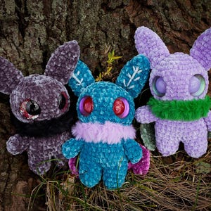 May include: Three crocheted plush toys in the shape of bunnies with large eyes. The bunnies are purple, teal, and black. The teal bunny has a purple fur collar and snowflake designs on its ears. The purple bunny has a green fur scarf.