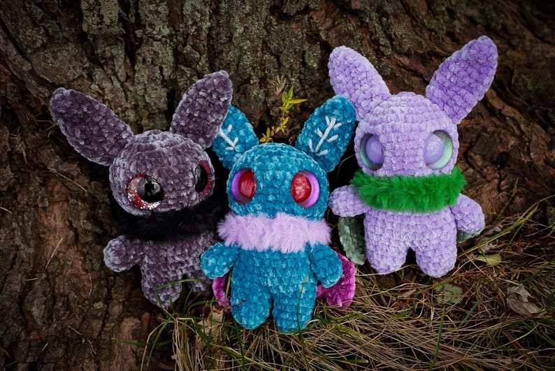 May include: Three crocheted plush toys in the shape of bunnies with large eyes. The bunnies are purple, teal, and black. The teal bunny has a purple fur collar and snowflake designs on its ears. The purple bunny has a green fur scarf.