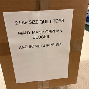 May include: A brown cardboard box with a white label. The label reads "2 LAP SIZE QUILT TOPS, MANY MANY ORPHAN BLOCKS, AND SOME SURPRISES". The box is sealed with tape and sits on a patterned surface.