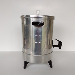 May include: A silver metal beverage dispenser with a black spigot and three black legs. The dispenser has a measurement scale on the side.