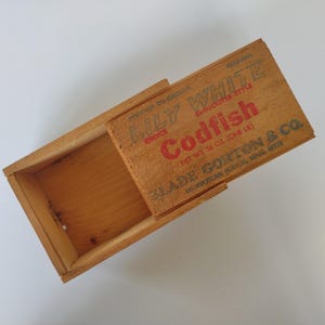 May include: A vintage wooden box with a sliding lid. The lid is printed with "LILY WHITE Codfish" in red and black text. The box is made of light-colored wood and appears to be an antique.