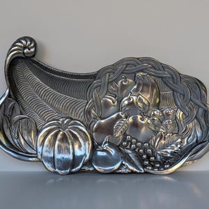 Puede incluir: Una bandeja decorativa plateada en forma de cuerno de la abundancia. La bandeja está llena de tallas en relieve detalladas de una calabaza, manzanas, peras, uvas y trigo. El diseño tiene un borde tejido.