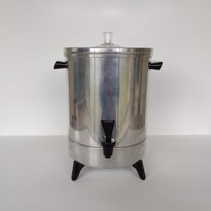 May include: A silver metal coffee pot with a black plastic spout and handles. The pot has a glass lid and sits on three black legs.