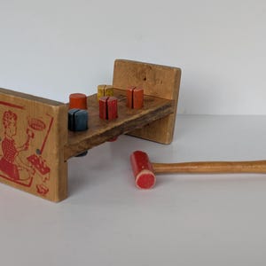 May include: A vintage wooden pounding bench toy with a red and white illustration on the side. The toy has a wooden hammer with a red head and a series of colorful pegs. The pegs are red, yellow, orange, and blue.