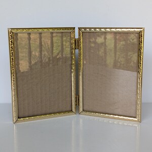 May include: A double hinged picture frame with a metallic gold finish and a textured, rope-like border pattern. The two rectangular frames are connected by a central metal hinge, allowing the unit to stand upright in an open, angled position.