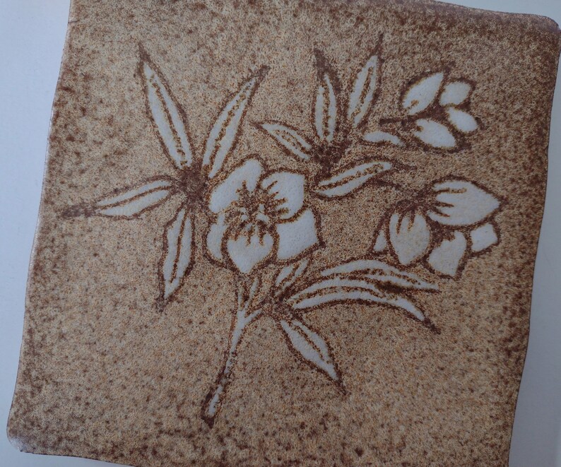 May include: Square ceramic tile with a floral design. The tile has a speckled, tan background with a brown outline of a flower and leaves. The flower petals are white. The tile is likely for home decor.