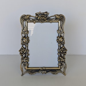 May include: Ornate bronze-colored picture frame with a rectangular opening. The frame is decorated with detailed rose and leaf motifs. The design features roses at the top and along the sides, with delicate leaf accents. The frame is suitable for displaying a photograph or artwork.