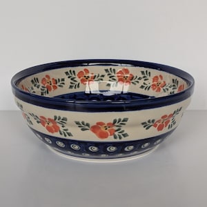 Op de afbeelding: Een ronde keramische kom met een crèmekleurige basis voorzien van een handgeschilderd patroon van oranje bloemen met groene bladeren. De rand en de voet zijn versierd met een donkerblauwe band en het onderste gedeelte bevat een blauw cirkelmotief.