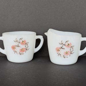 May include: White milk pitcher and sugar bowl set. Each piece features a floral design with pink and orange flowers and brown stems. The sugar bowl has two handles, while the pitcher has a handle and a spout.