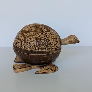 Könnte beinhalten: Eine braune Schildkrötenfigur aus Holz mit einem abgerundeten Panzer, der mit filigranen, hellen Gravuren von Spiralen und echsenartigen Formen verziert ist. Die Skulptur hat eine glatte, dunkelbraune Oberfläche und flach geschnitzte Füße.