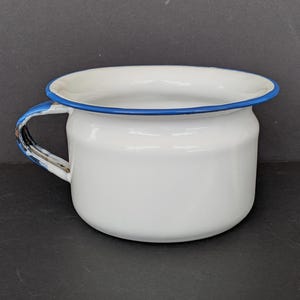 May include: A white enamelware mug with a blue rim and handle. The mug has a rounded shape and a sturdy handle. The blue paint on the handle shows signs of wear, adding to its vintage charm. This mug is perfect for camping or rustic decor.