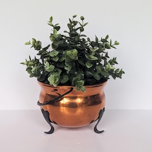 Puede incluir: Una maceta de metal color cobre brillante con cuerpo redondeado, asa negra y tres patas negras curvadas. La maceta contiene una planta artificial verde y frondosa con pequeñas hojas redondeadas que llenan toda la abertura superior.