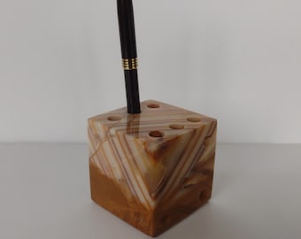 May include: A cube-shaped pen holder made of layered, light brown and cream-colored stone. A black pen with gold accents is inserted into one of the holes. The pen holder has several holes for pens or pencils.
