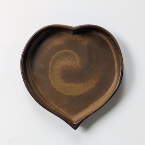 Può includere: Un piatto in ceramica a forma di cuore con esterno marrone scuro e interno marrone più chiaro e maculato, caratterizzato da un sottile motivo a spirale. Il piatto presenta un bordo liscio leggermente rialzato e una base piatta color terra.