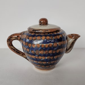 Op de afbeelding: Een klein, bruin en blauw aardewerken theepot met een deksel. De theepot heeft een gespikkeld ontwerp met bruine stippen op een blauwe achtergrond.