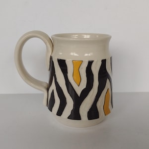 May include: A white ceramic mug with a black and yellow zebra stripe pattern. The mug has a rounded body and a handle.