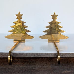 May include: A pair of polished brass stocking holders, each featuring a flat, stylized Christmas tree silhouette topped with a star. The base of each tree connects to a long, curved metal hook designed to hang over the edge of a mantelpiece.