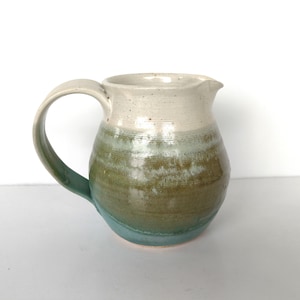 May include: A handmade ceramic pitcher with a white, green, and brown glaze. The pitcher has a rounded body and a handle.