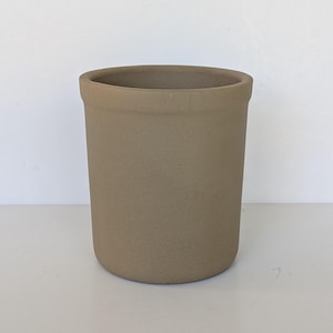 May include: A cylindrical stoneware crock with a matte, earthy taupe finish. The vessel features a simple, straight-sided design with a slightly thickened, rounded rim at the top and a flat base, presenting a uniform, solid-colored exterior surface.
