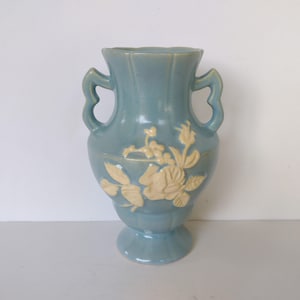 May include: A light blue ceramic vase with two handles and a white floral design. The vase has a classic, elegant shape.