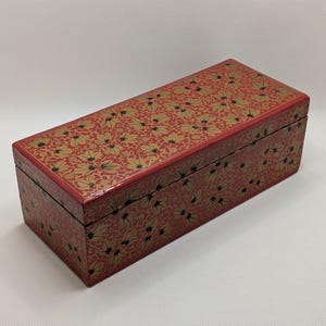 May include: A rectangular red box with a floral pattern in gold and black. The box has a glossy finish and a hinged lid. The floral design covers the top and sides of the box, creating a decorative storage container.