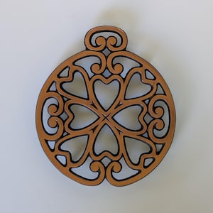 May include: A circular tan trivet featuring a symmetrical openwork design of interconnected heart shapes and swirling scroll patterns. The object has a smooth, flat surface with dark brown edges outlining the intricate cutouts throughout the piece.