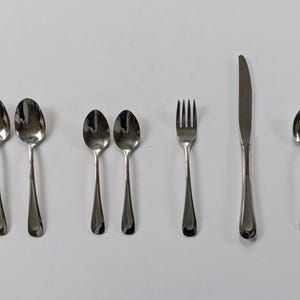 Könnte beinhalten: Ein Set aus Edelstahlbesteck, darunter Löffel, eine Gabel und ein Messer. Die Löffel haben ovale Schalen und lange, schlanke Griffe. Die Gabel hat vier Zinken und das Messer hat eine gebogene Klinge. Das Besteck ist auf einer weißen Oberfläche angeordnet.