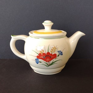 May include: A white teapot with a yellow lid and a floral design featuring a red poppy, blue flowers, and green leaves.