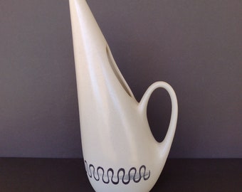 May include: A white ceramic jug with a distinctive, angular design and a black wavy line pattern around the base. The jug has a spout and a handle.