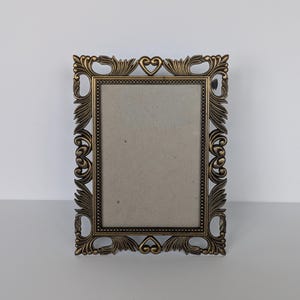 May include: Ornate rectangular picture frame with a bronze finish. The frame features intricate floral and heart-shaped detailing around the edges. The center holds a neutral-colored backing, ready for a photograph or artwork. A decorative piece for displaying memories.