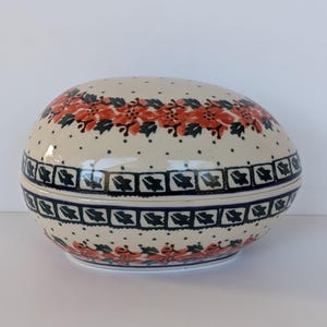 May include: A ceramic egg-shaped trinket box with a lid. The box is cream-colored with a floral pattern in red and green. A band of dark blue and black geometric shapes encircles the middle. Small black dots are scattered across the surface.