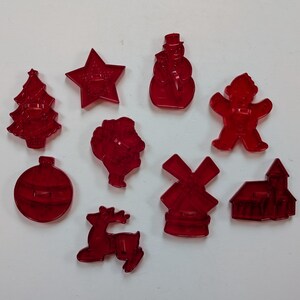 May include: A collection of nine red plastic cookie cutters in various shapes including a Christmas tree, a star, a snowman, a gingerbread person, a round ornament, a Santa Claus figure, a windmill, a reindeer, and a small house structure.