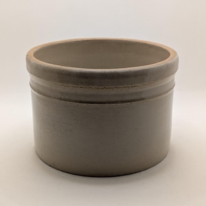 May include: A gray stoneware crock with a cylindrical shape. The crock has a slightly flared rim and a band of raised detail around the middle. The interior is a lighter shade of gray. The piece has a rustic, vintage aesthetic.