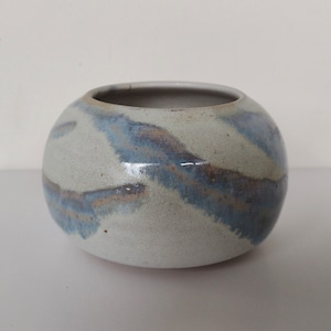 May include: A small, round ceramic vase with a white glaze and blue and brown streaks. The vase is perfect for displaying small flowers or plants.