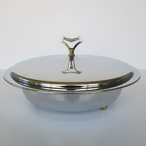 Puede incluir: Una fuente ovalada plateada con tapa a juego y asa decorativa. La fuente tiene un borde elevado y se apoya en tres pequeños pies dorados. El asa de la tapa tiene un diseño estilizado único.