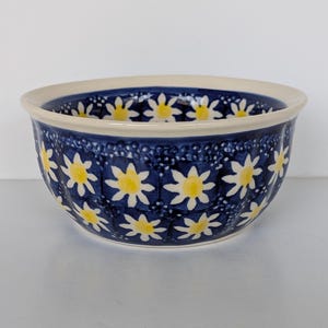 May include: A ceramic bowl with a dark blue background and a pattern of yellow and white daisy-like flowers. The bowl has a cream-colored rim and a rounded shape. The floral design is evenly spaced around the bowl's exterior.