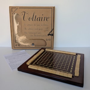 May include: A Voltaire solitaire word game set featuring a brown cardboard box with a clear window, a dark brown wooden game board with a grid and light-colored letter tiles, and a white instruction sheet with printed text in multiple languages.