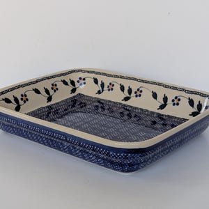 Pode incluir: Uma caneca de cerâmica branca com uma alça preta e o texto "But First, Coffee" em negrito preto. A caneca está sobre um fundo desfocado, sugerindo um ambiente acolhedor.