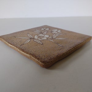 May include: A square, tan ceramic coaster with a floral design. The flowers and leaves are white, and the coaster has a slightly textured surface. The edges are a darker brown.